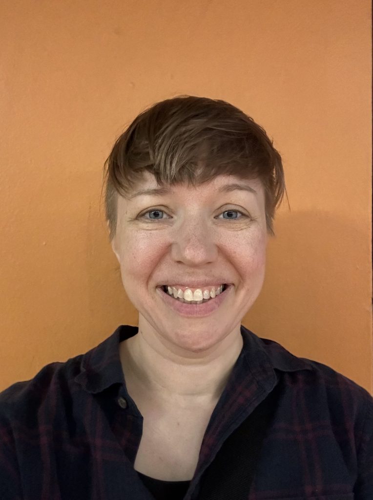 A smiling person with short hair, wearing a plaid shirt, standing in front of an orange wall.